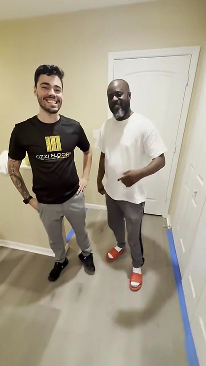 Someone from Ozzifloors with a homeowner in a hallway, the new grey plank down and painter’s tape still along the baseboard